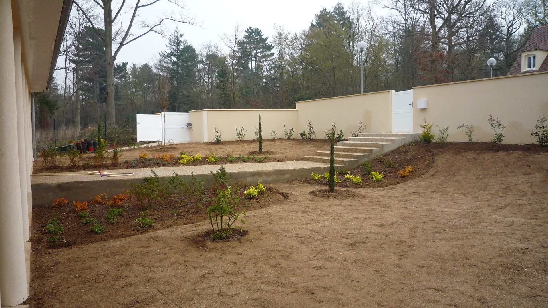 Un jardin en terrasses à l'arrière de la maison, avec des murs de soutènement couleur crème, des escaliers, de jeunes plantations et de la terre nue sous un ciel nuageux.