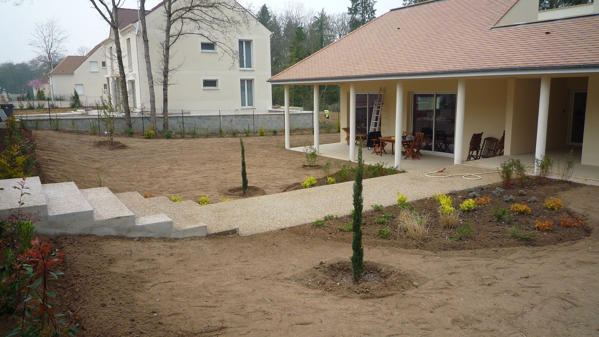 Une maison moderne aux tons clairs, avec un toit de tuiles rouges, une cour gravillonnée, des marches en pierre et un chemin de jardin paysager.