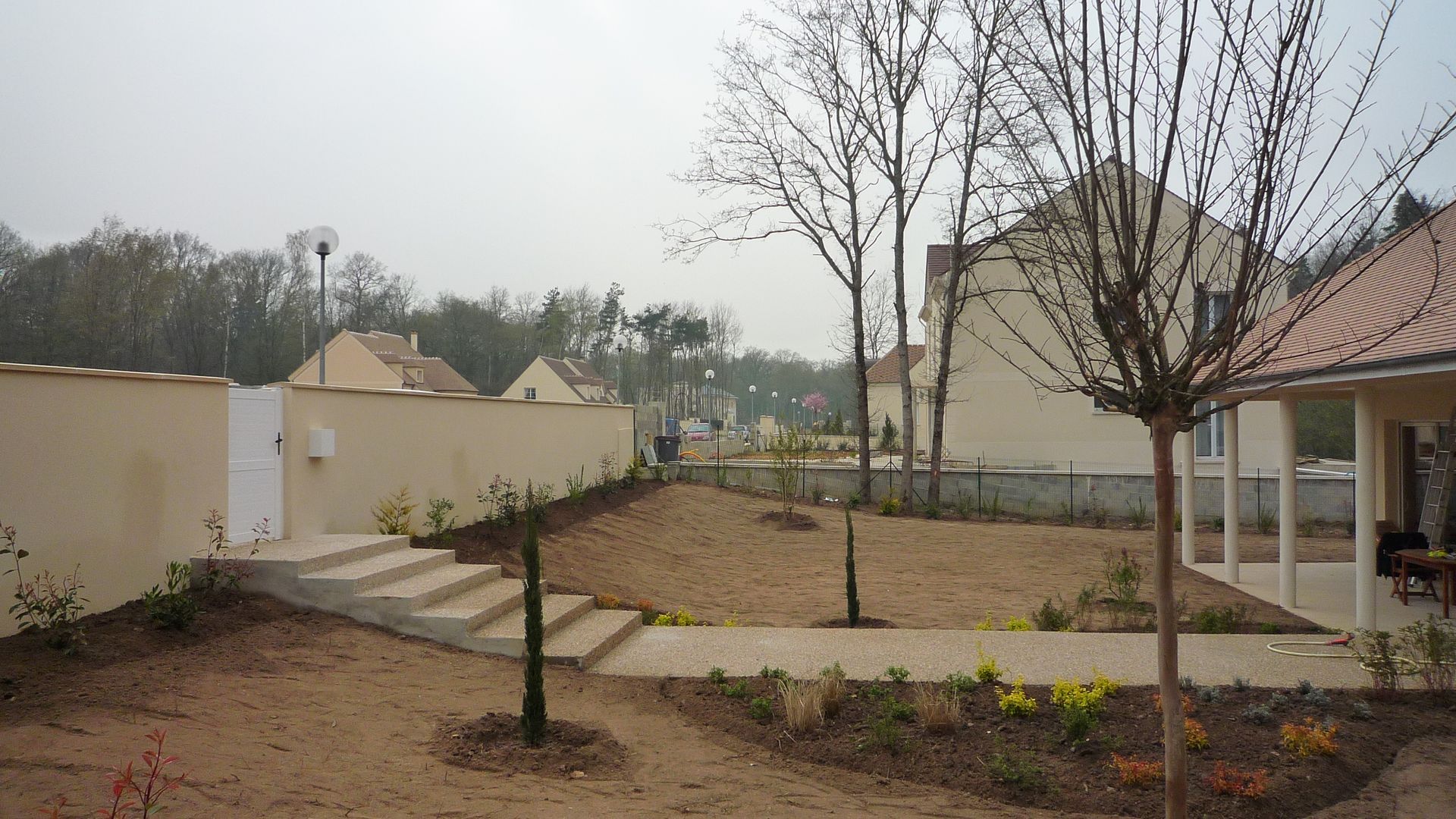 Un mur beige avec un portail et des marches en pierre mène à un jardin arrière arboré, agrémenté d'un chemin pavé et de nouvelles plantations.