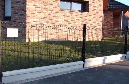 Une clôture en grillage noir se dresse sur un trottoir en béton bas, devant une maison en briques rouges entourée d'une pelouse verte.