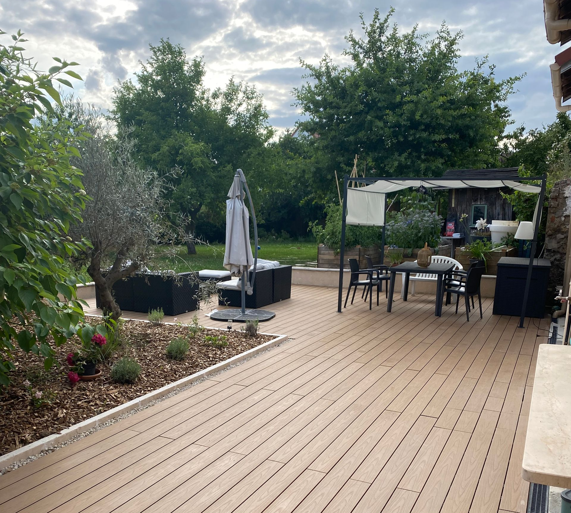 Terrasse avec mobilier de jardin, arbres et ciel nuageux.