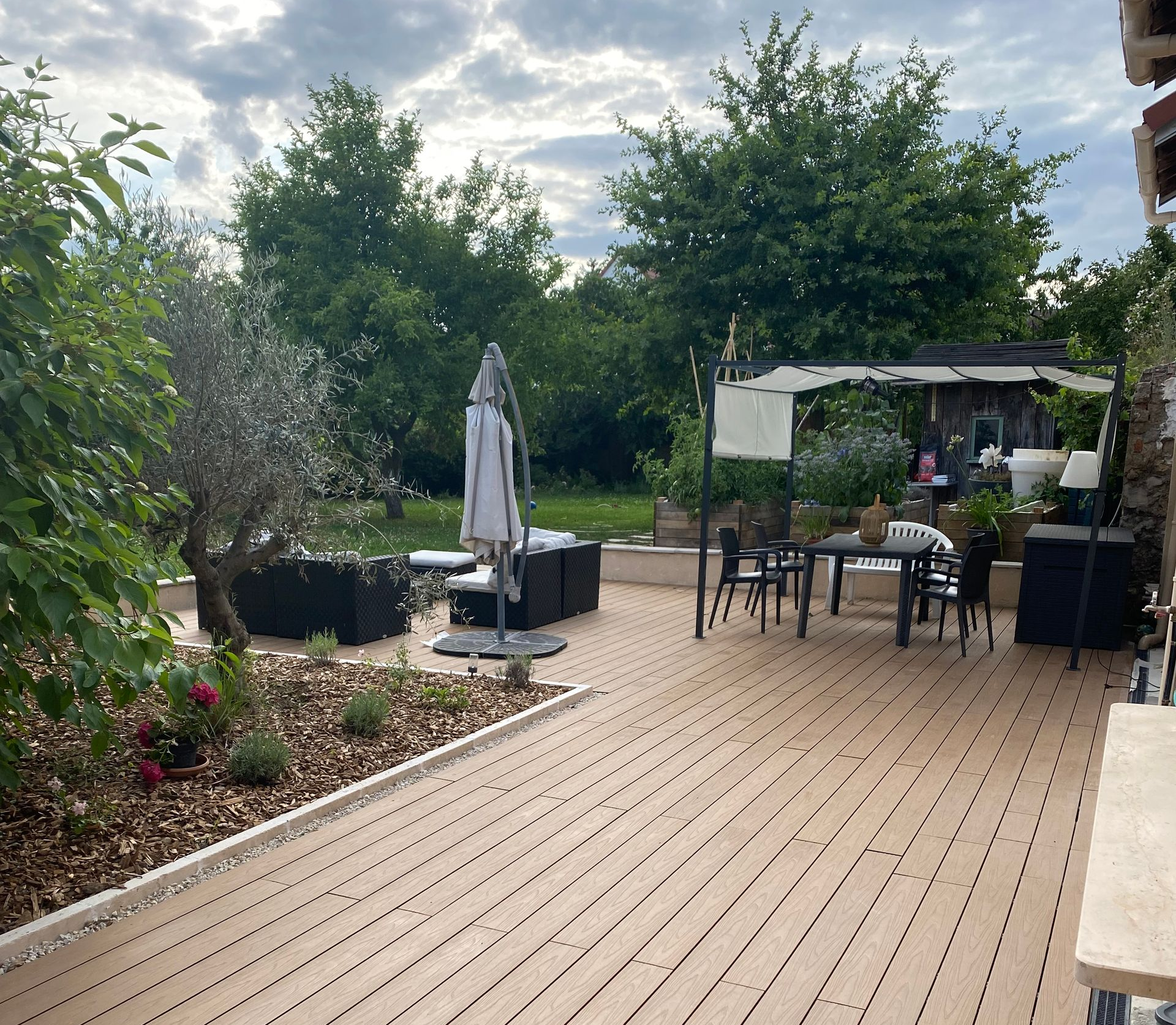 Terrasse en bois avec mobilier de jardin, parasol et jardin arboré en arrière-plan. Ciel nuageux.