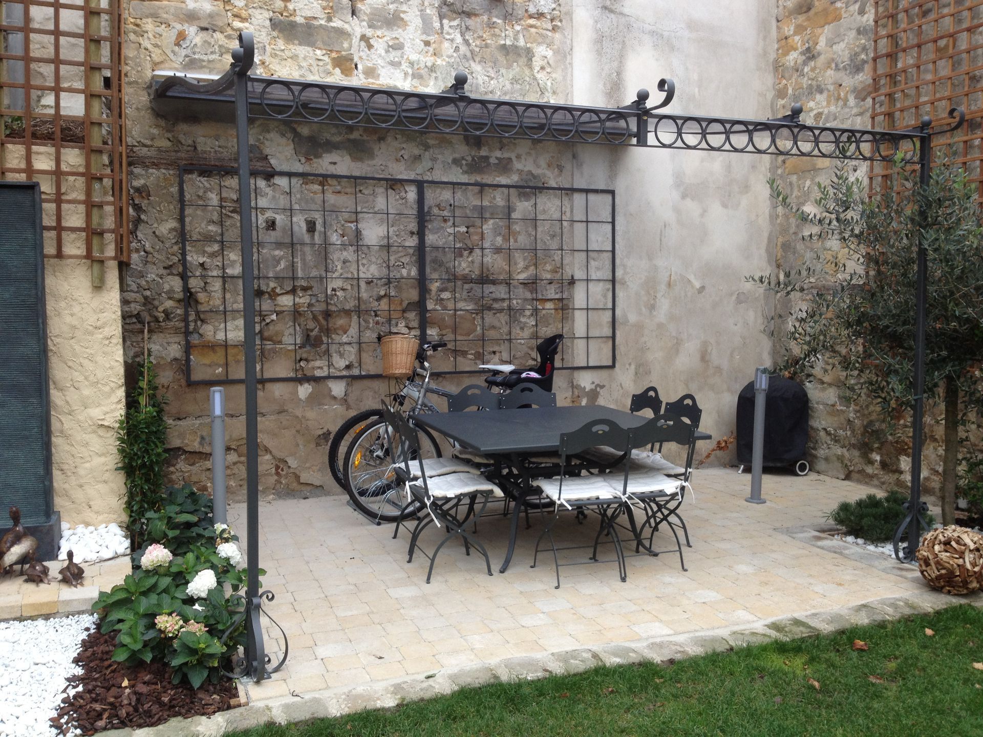 Terrasse avec table, chaises et vélo sous une pergola en fer forgé devant un mur en pierre.