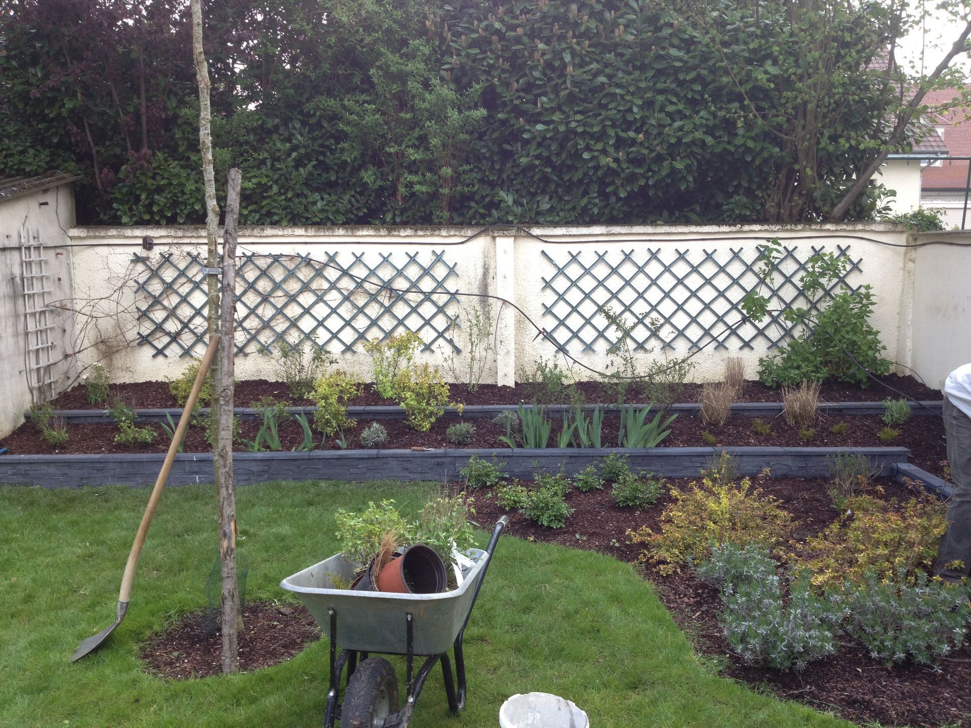 Un parterre de jardin avec une brouette, une pelle et un treillis adossé à un mur. On aperçoit du paillis et des plantes.