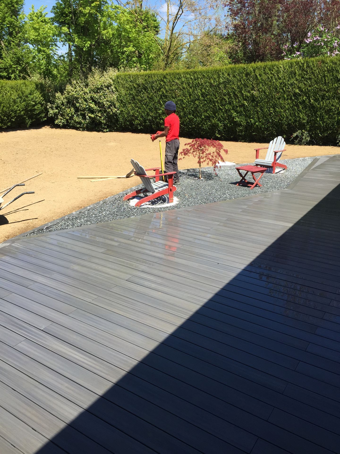 Une personne vêtue d'un t-shirt rouge se trouve sur une terrasse en bois, en train d'utiliser un outil. Deux chaises rouges sur de petites estrades sont disposées à proximité.
