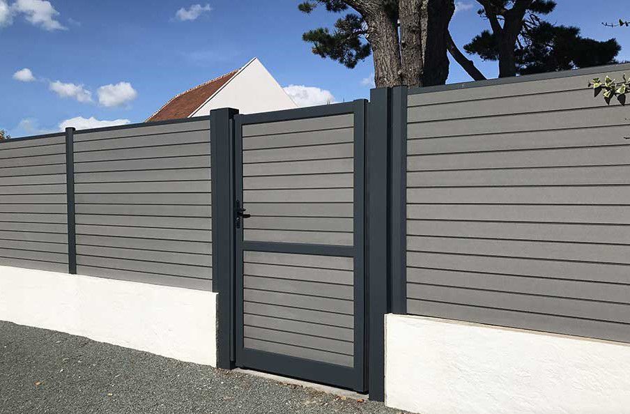 Clôture à lames horizontales avec portail assorti devant un bâtiment blanc, sous un ciel bleu.