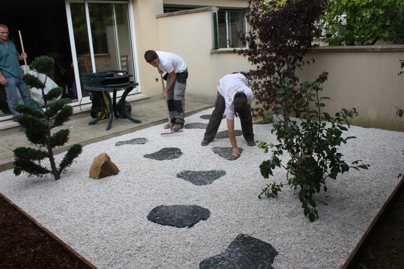 Deux personnes disposent des dalles de jardin dans une cour recouverte de gravier, avec des plantes et une personne à proximité.