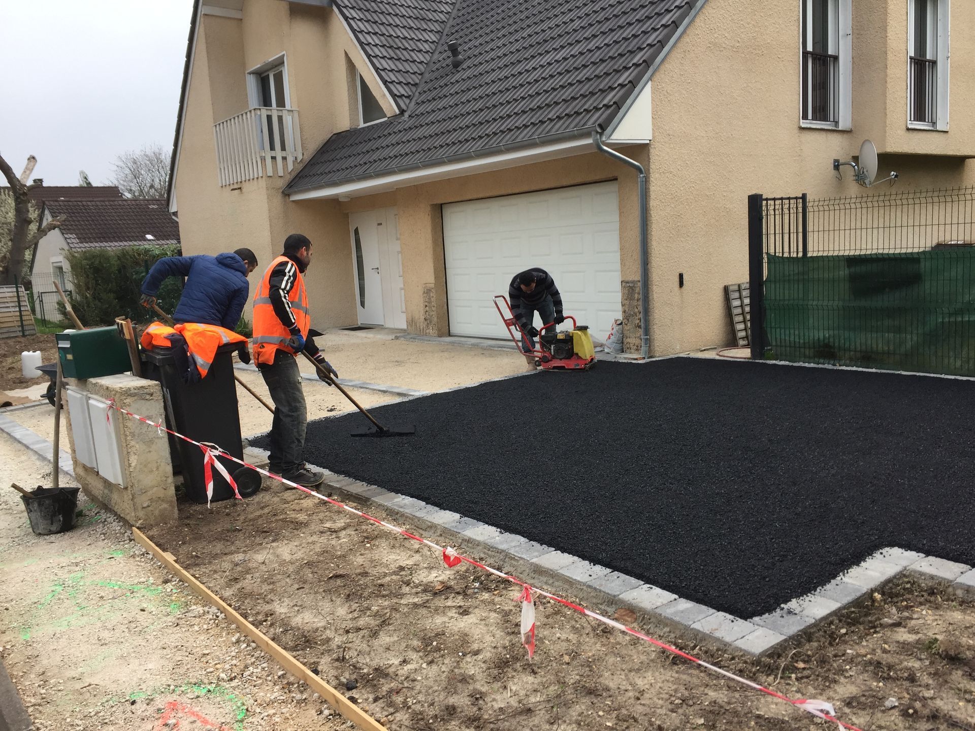 Des ouvriers asphaltent une allée. Une maison beige avec garage est visible en arrière-plan. Un ouvrier utilise un compacteur.