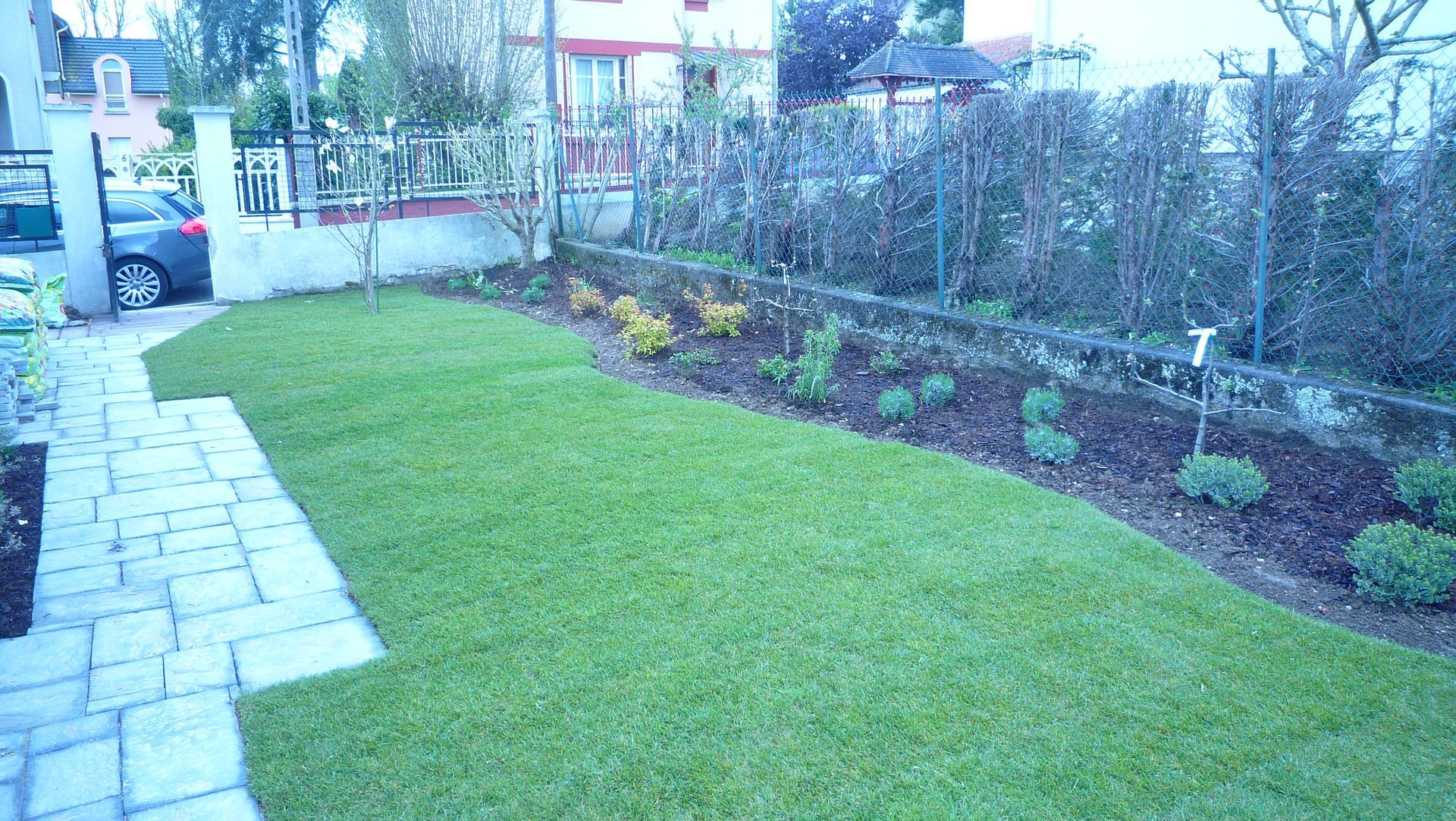 Pelouse et parterre de fleurs dans le jardin de devant, avec un chemin de pierre, une voiture garée à proximité et une haie en arrière-plan.