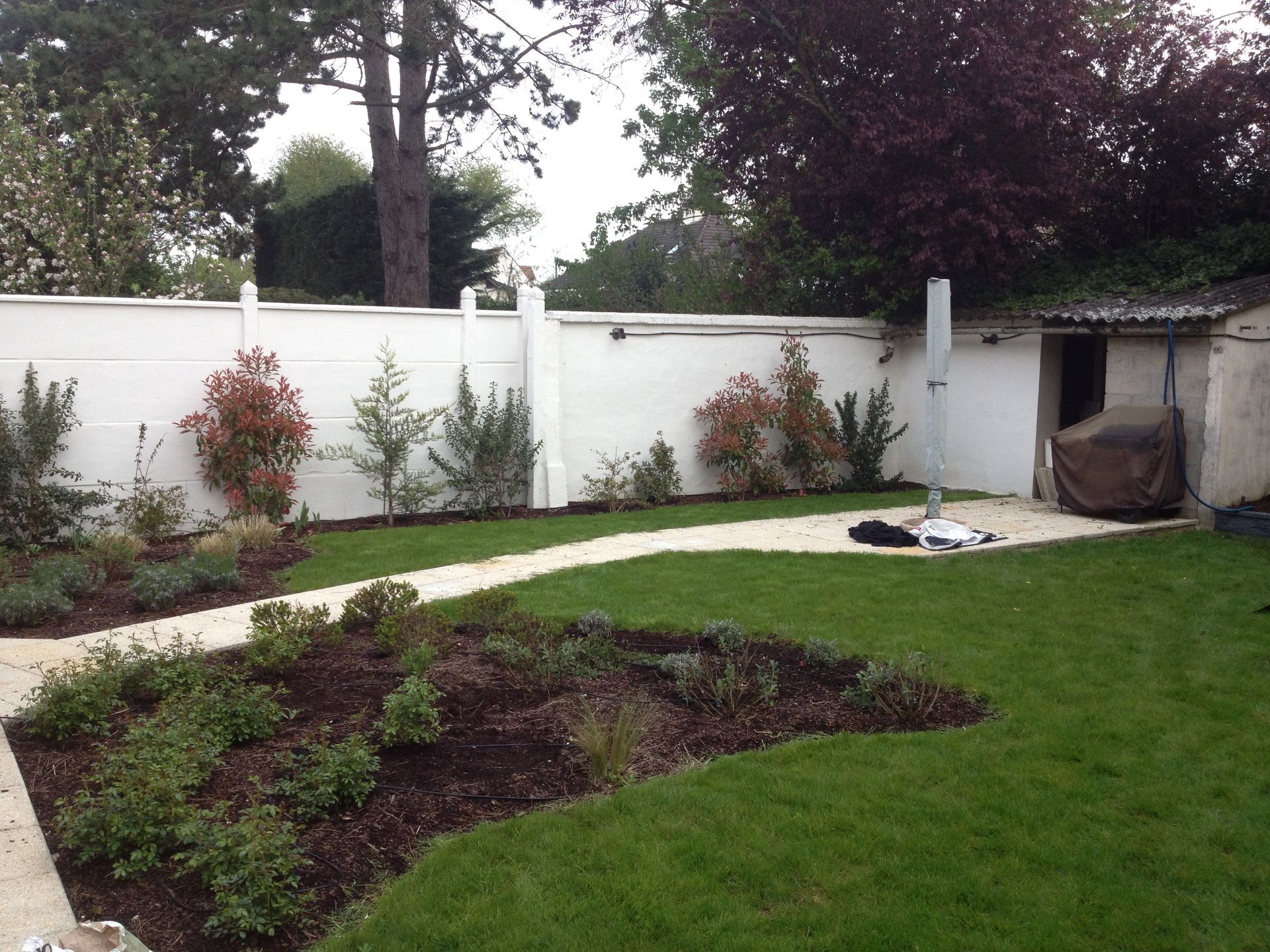 Jardin avec allée en pierre, pelouse et parterres de fleurs adossés à un mur blanc et un abri de jardin.