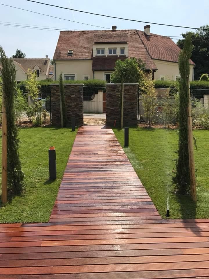 Une allée en bois mène à une entrée bordée d'herbe et d'arbres, une maison se dressant en arrière-plan.