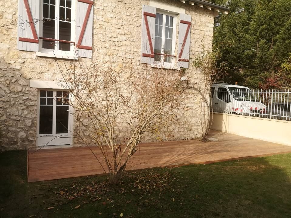 Maison en pierre avec terrasse en bois, petit arbre et véhicule blanc garé derrière une clôture.