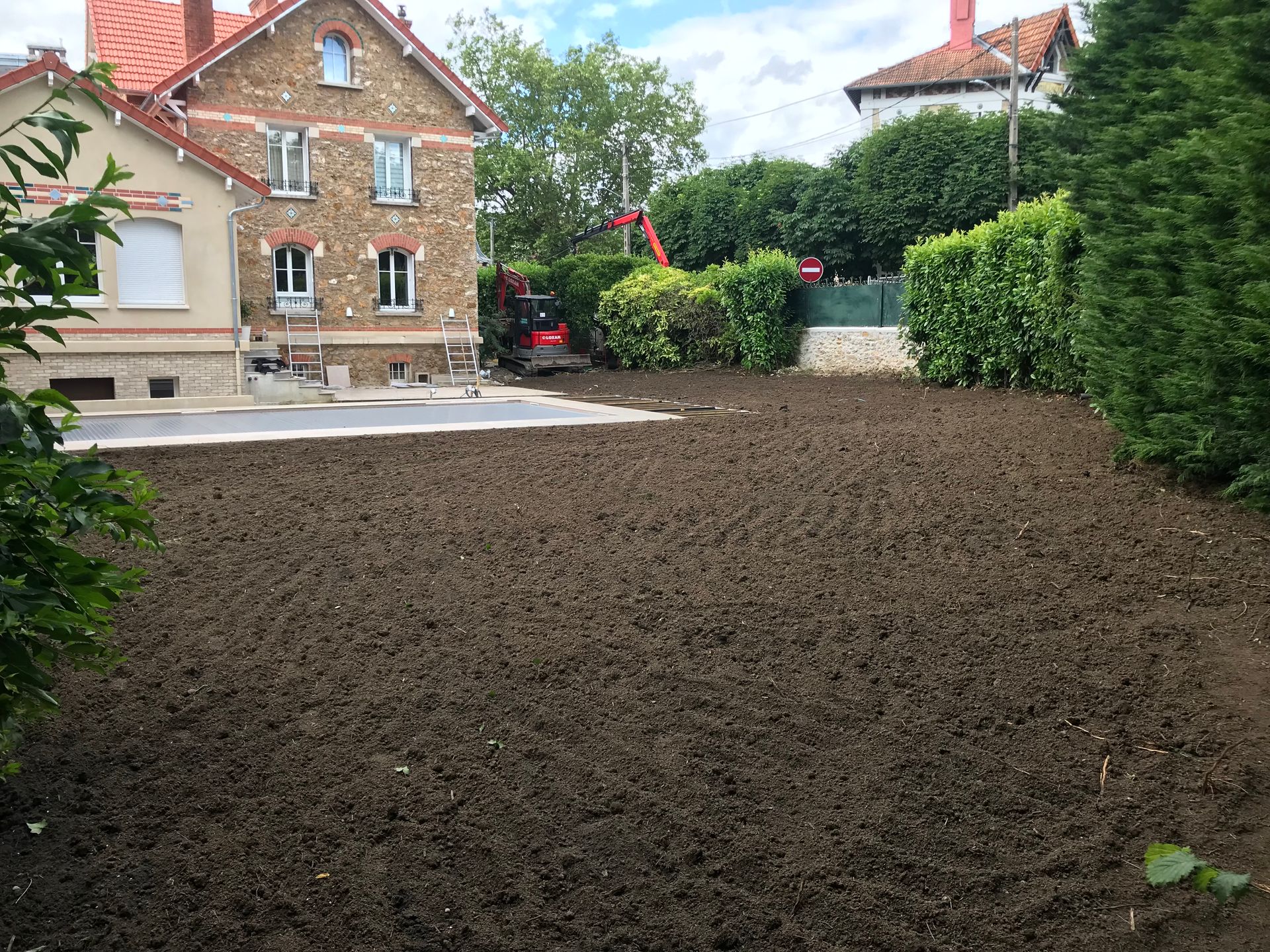 Pelouse recouverte de terre devant une maison en briques ; engins de chantier en arrière-plan.