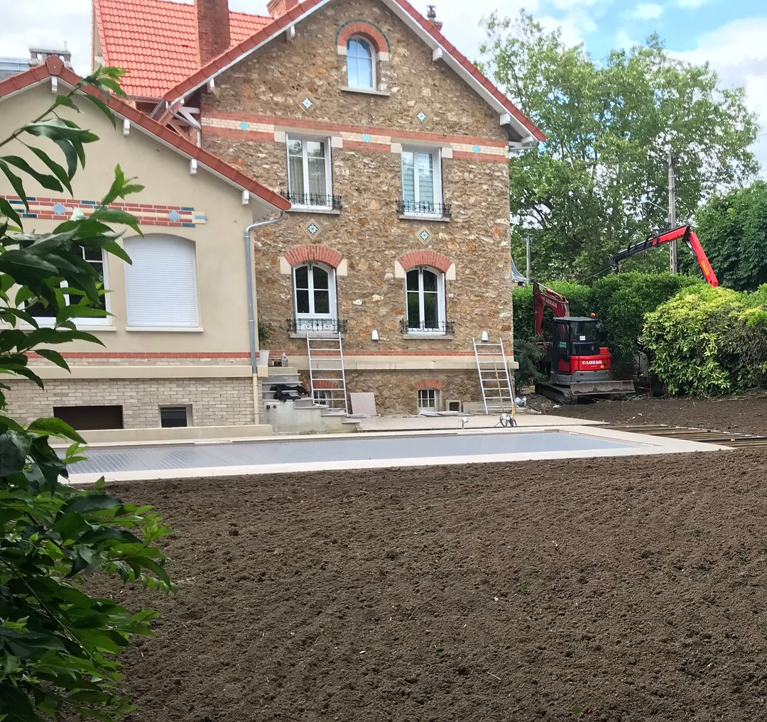 Un jardin avec un sol préparé devant une maison en briques. Des arbustes verts bordent le jardin.