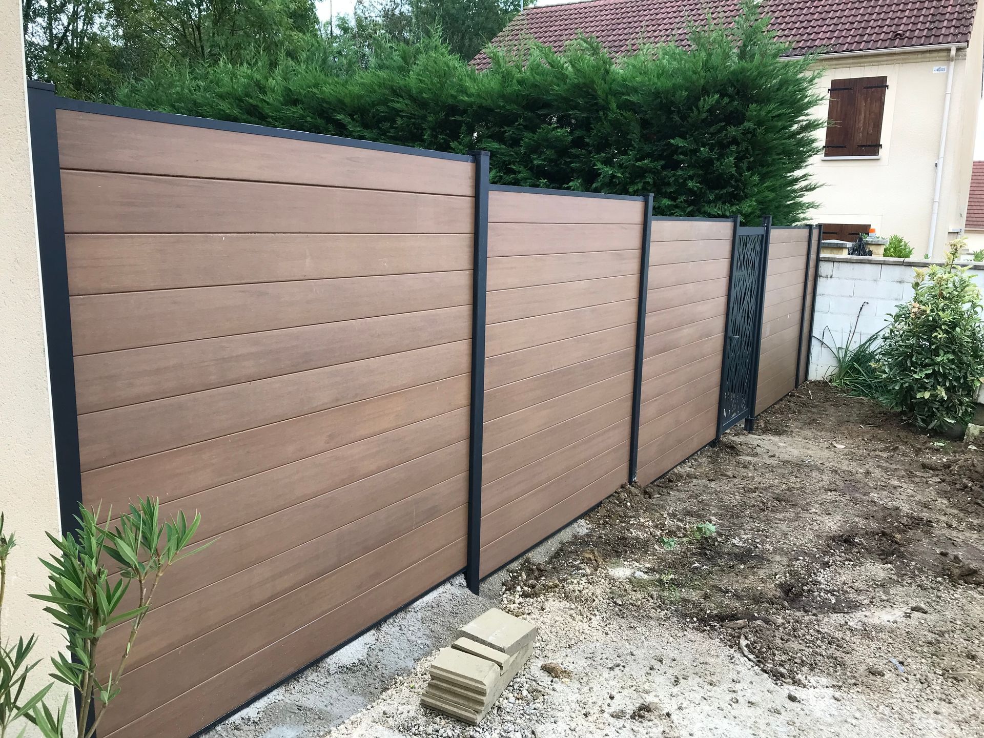 Clôture composite marron avec poteaux en métal noir, donnant sur une cour partiellement aménagée avec des haies vertes en arrière-plan.