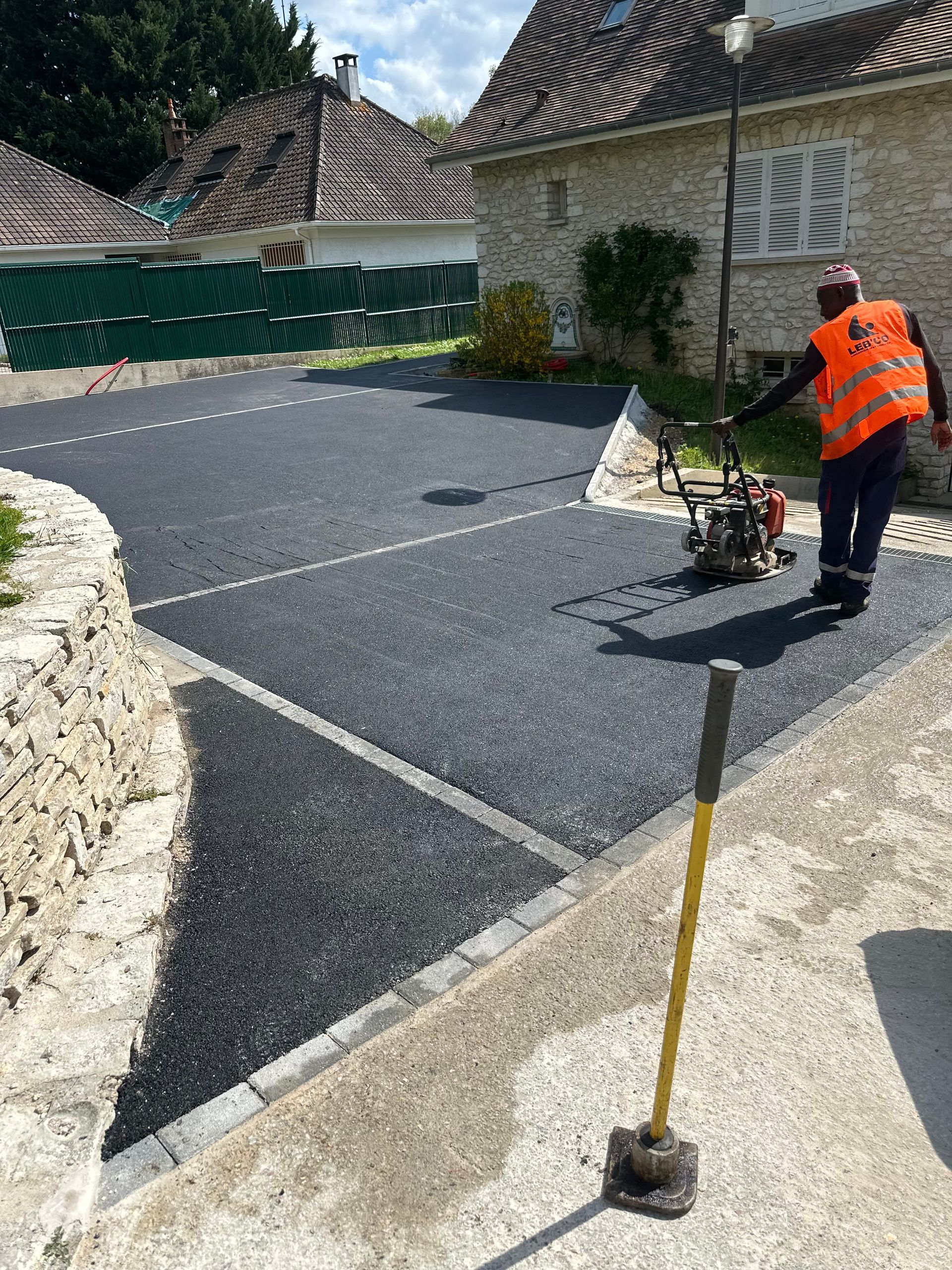 Un ouvrier portant un gilet de sécurité compacte de l'asphalte frais sur une surface pavée près d'un bâtiment.