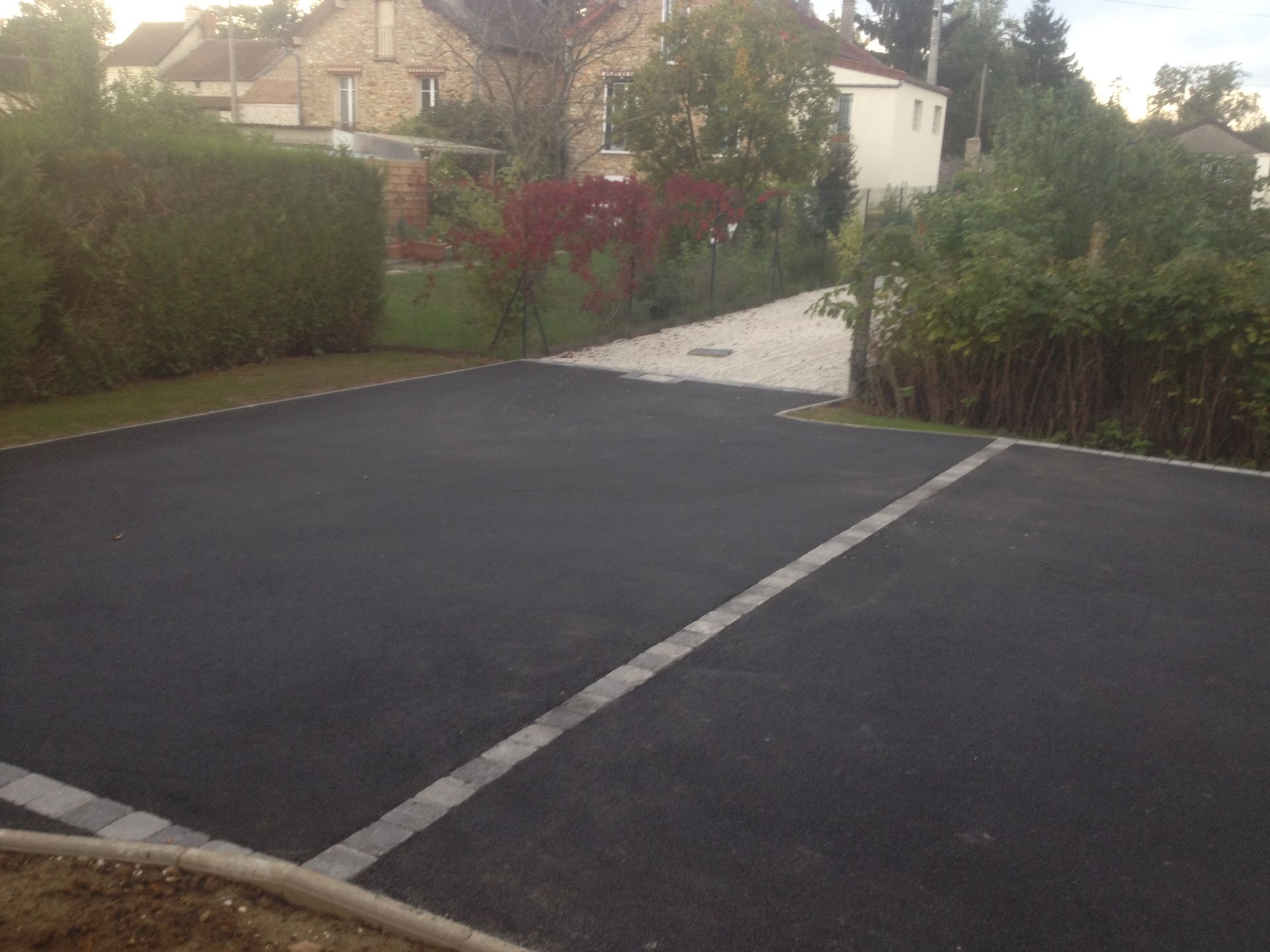 Allée en asphalte noir avec ligne blanche, bordée de haies et de verdure, menant à un chemin de couleur claire.