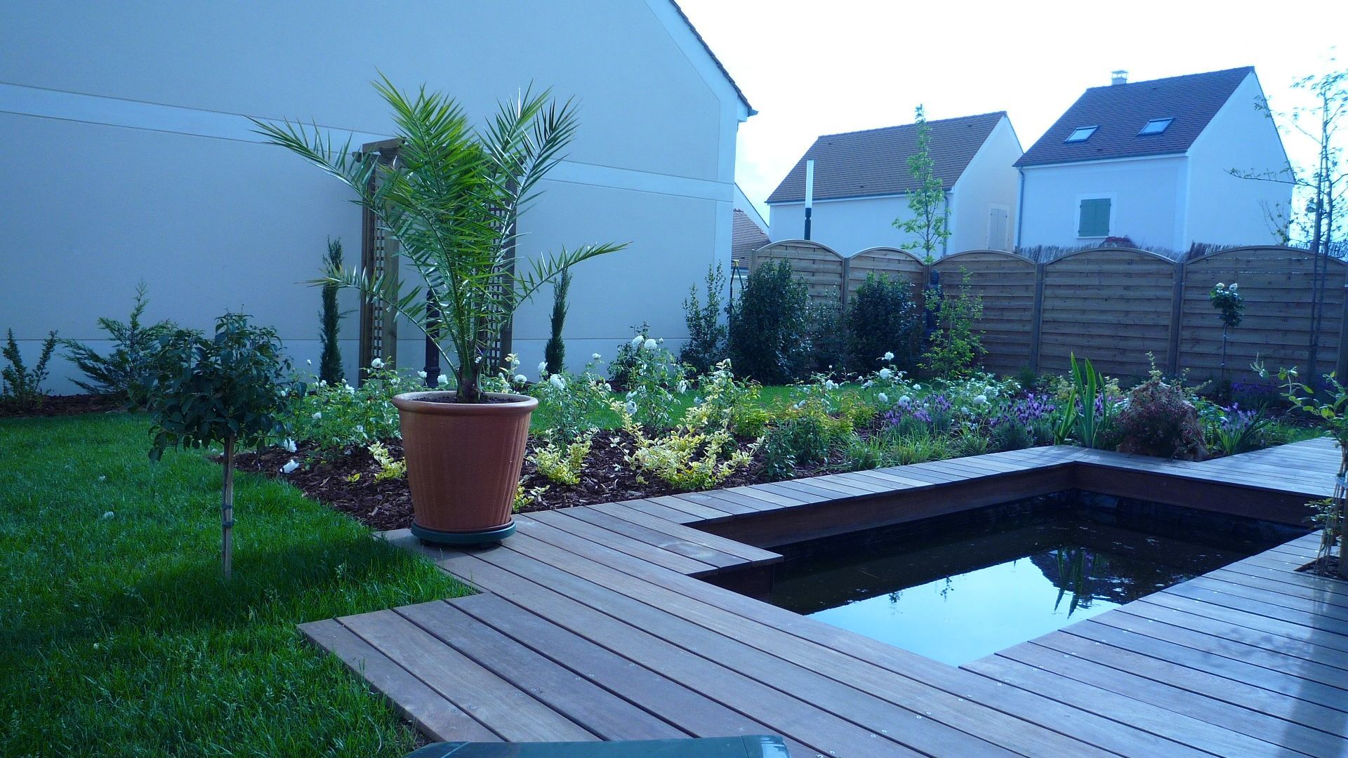 Jardin avec terrasse en bois, petit bassin, palmier en pot et plantes diverses. Maisons en arrière-plan.