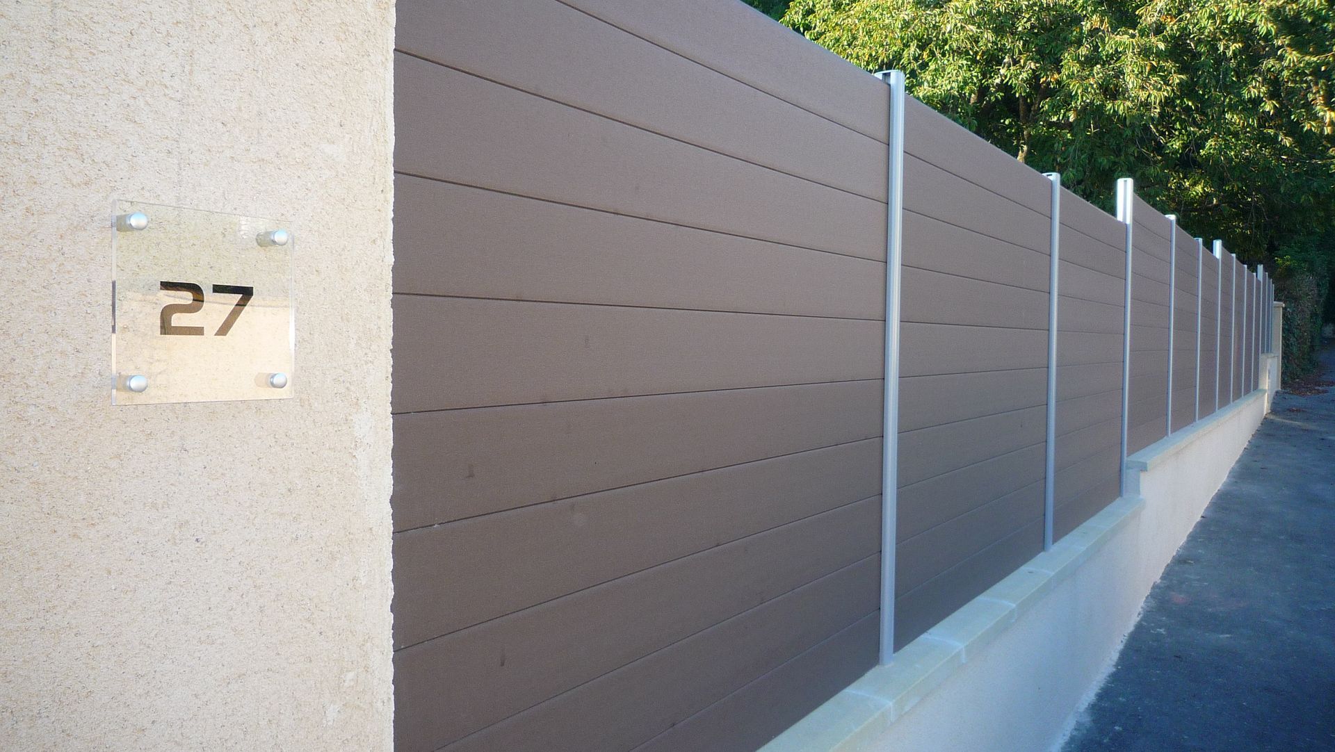 Mur en stuc gris avec un numéro de maison en métal. Clôture à lattes horizontales marron avec poteaux métalliques.