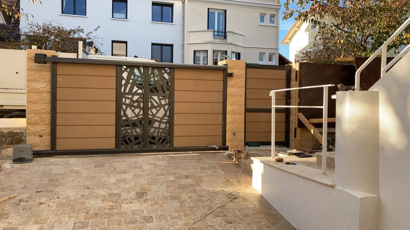 Portail en bois avec panneau central décoratif, colonnes en pierre beige et mur blanc adjacent.