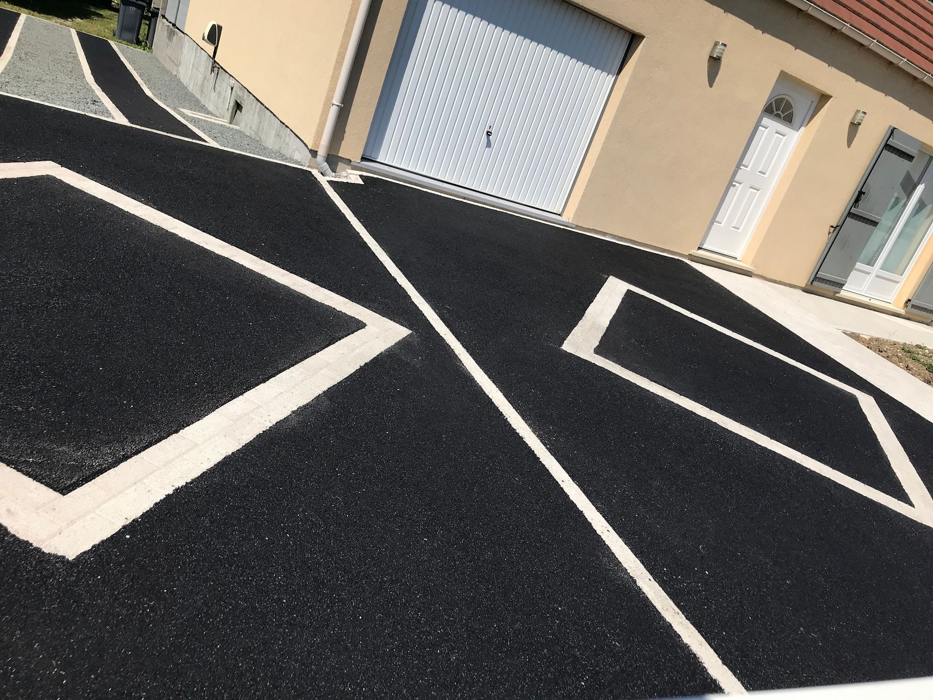 Allée en asphalte noir ornée de motifs géométriques blancs, menant à une maison beige avec garage.