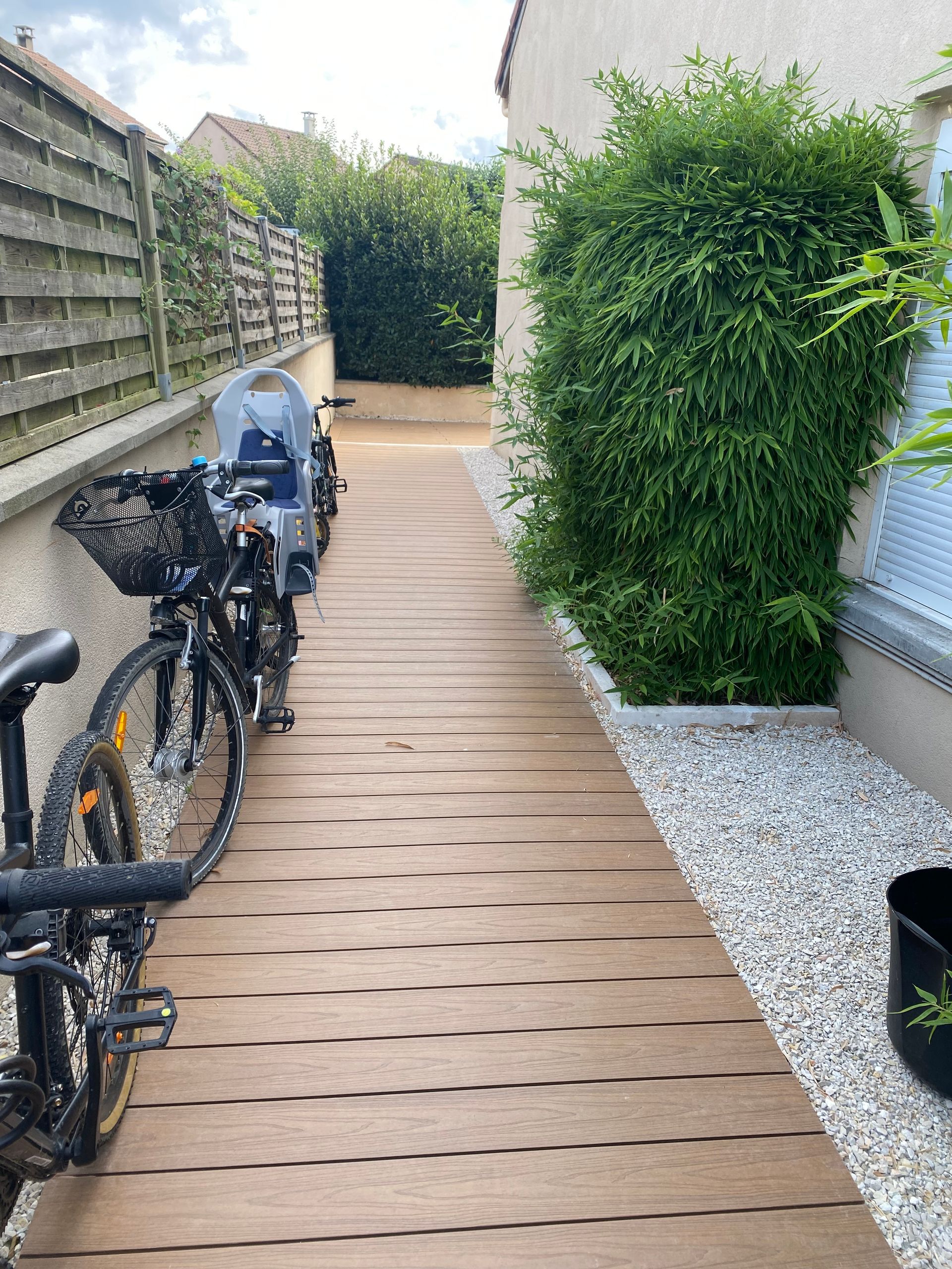 Allée en bois longeant un bâtiment, avec des vélos garés d'un côté et de la verdure de l'autre.