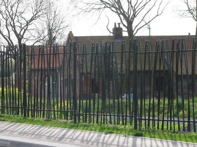 Clôture en barres noires devant un bâtiment en briques. Herbe et arbres en arrière-plan, journée ensoleillée.