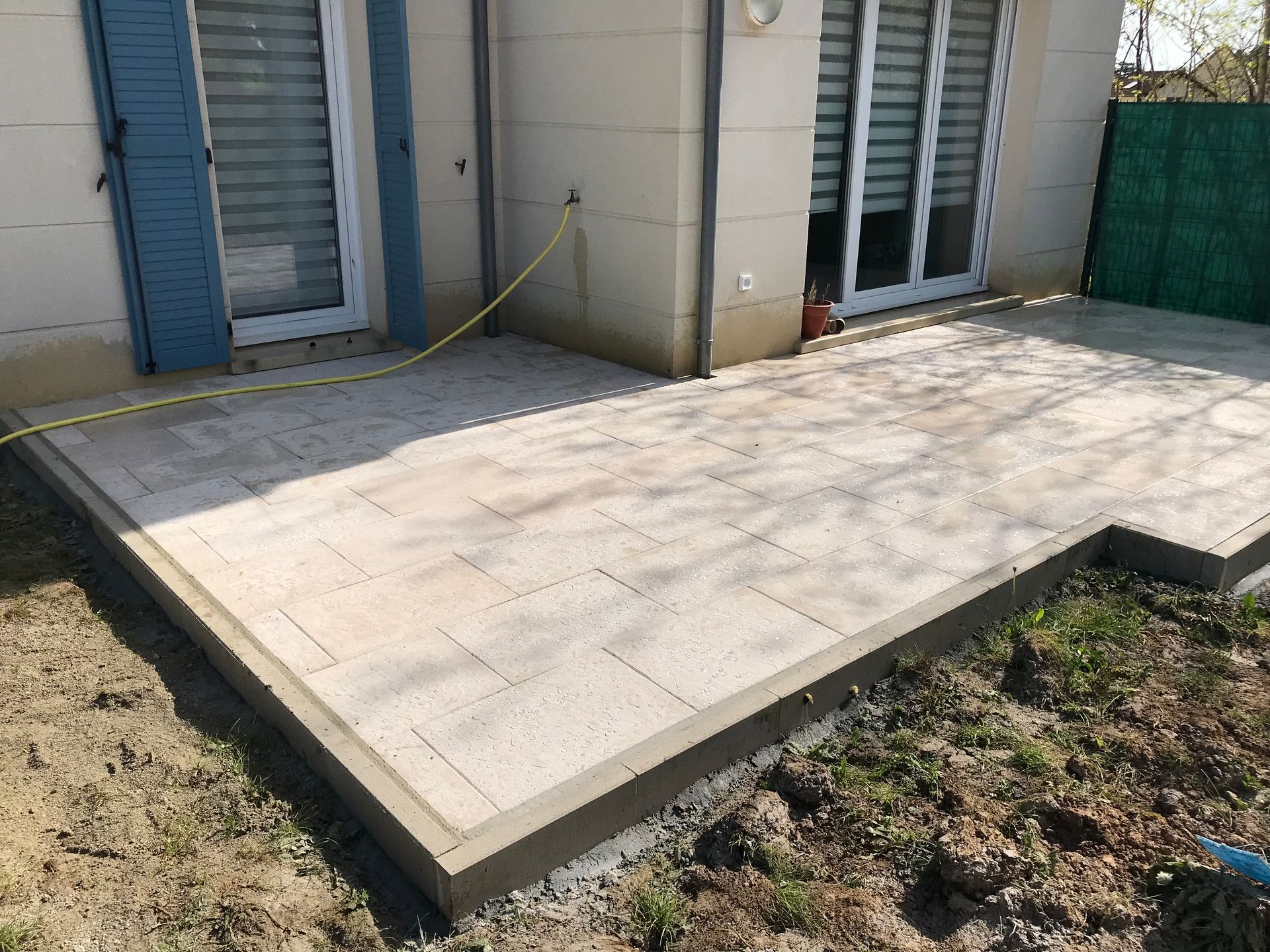Une terrasse pavée devant une maison. Des carreaux beiges bordés de gris, jouxtant la pelouse et un mur.