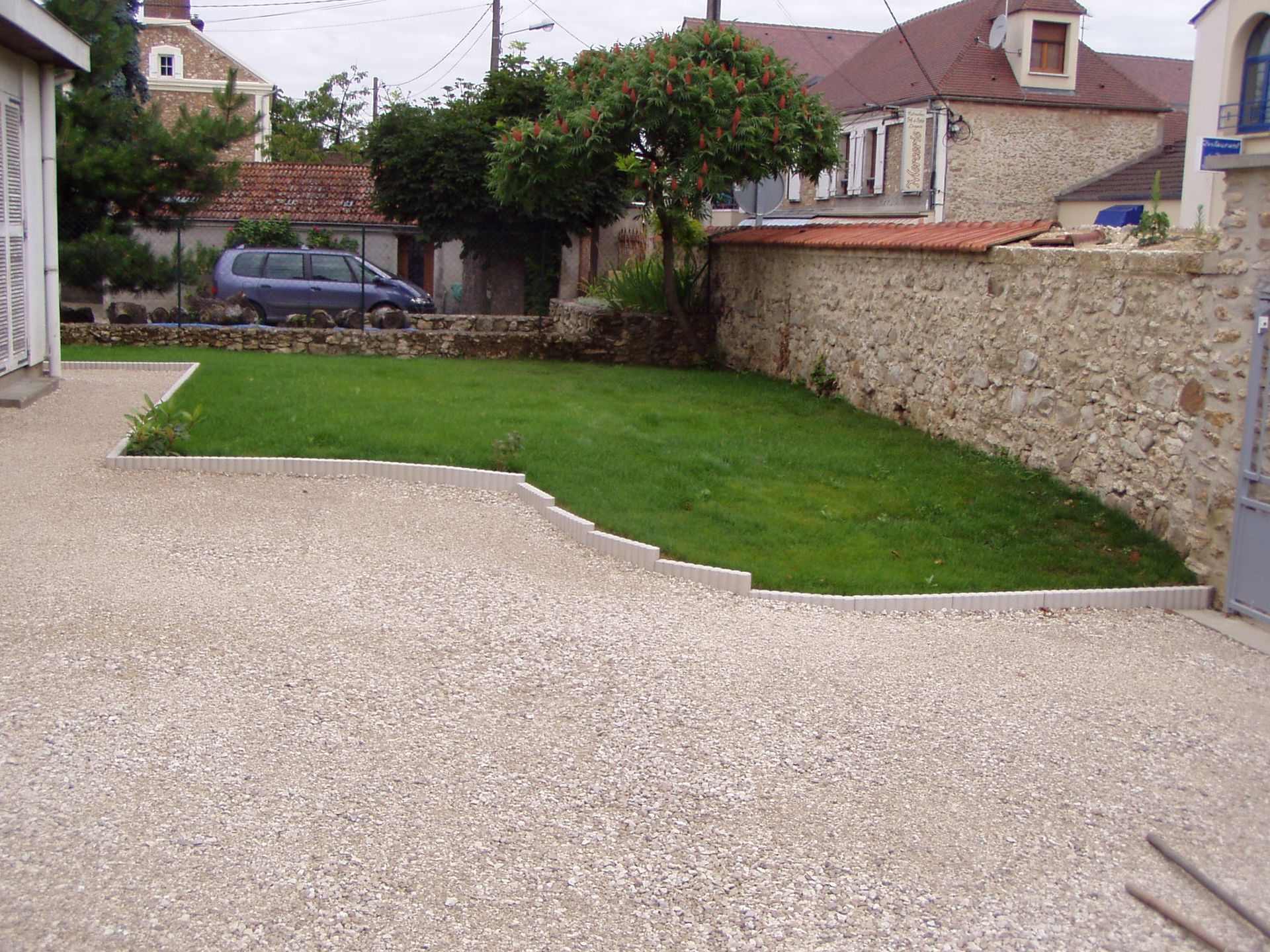Une allée de gravier mène à une petite pelouse verte bordée d'un muret de pierre. Des maisons se dressent à l'arrière-plan.