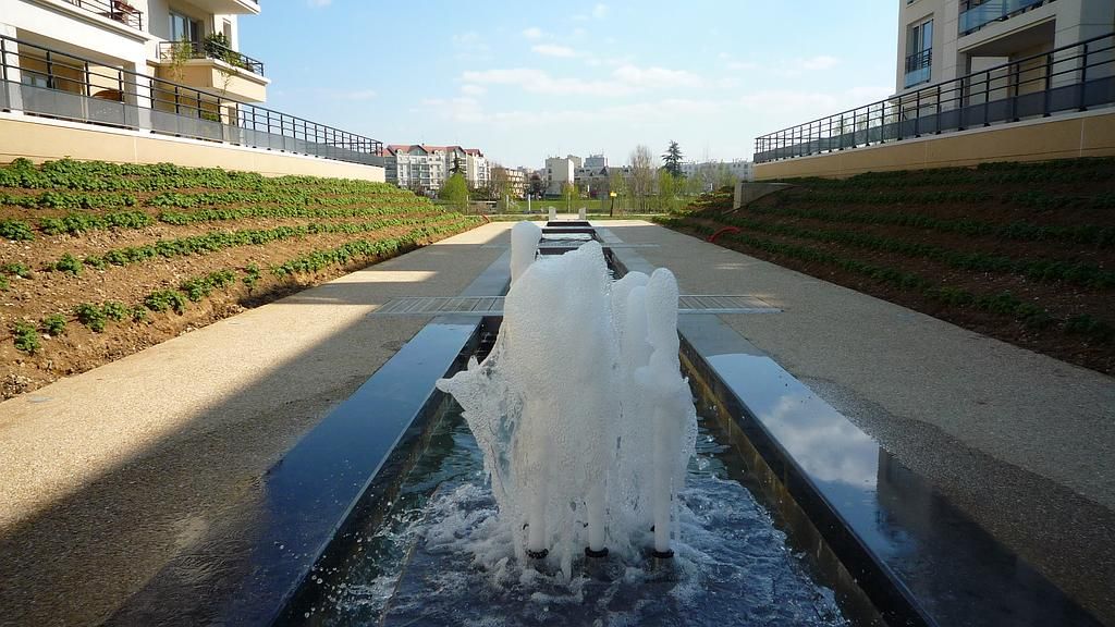 Une fontaine projette de l'eau vers le haut sur une place pavée, avec des immeubles d'appartements en arrière-plan. Journée ensoleillée.