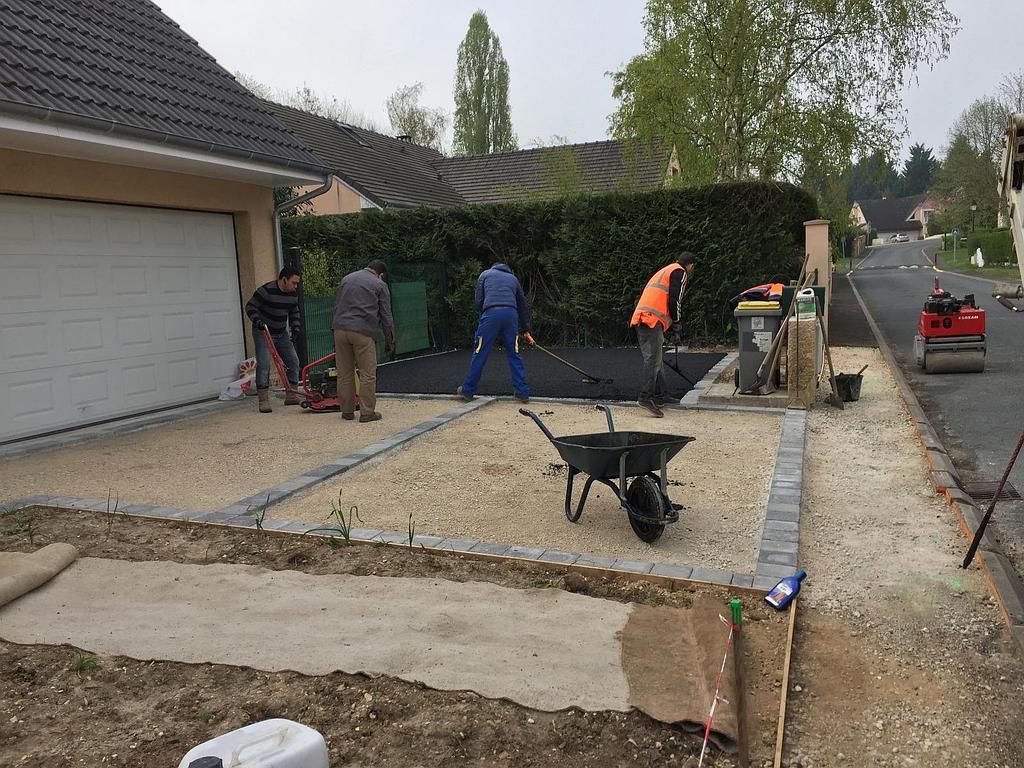 Des ouvriers réalisent le pavage d'une allée à l'aide d'asphalte et d'un rouleau compresseur. Bordures de gravier, haie verte, porte de garage.