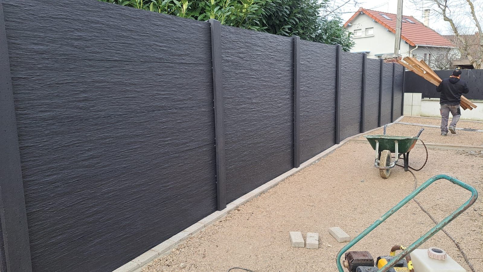 Une clôture en béton noir est en construction dans un jardin ; une personne transporte des planches de bois.