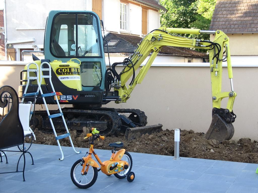Une pelleteuse jaune creuse la terre à côté d'un mur blanc, avec un vélo orange d'enfant devant.
