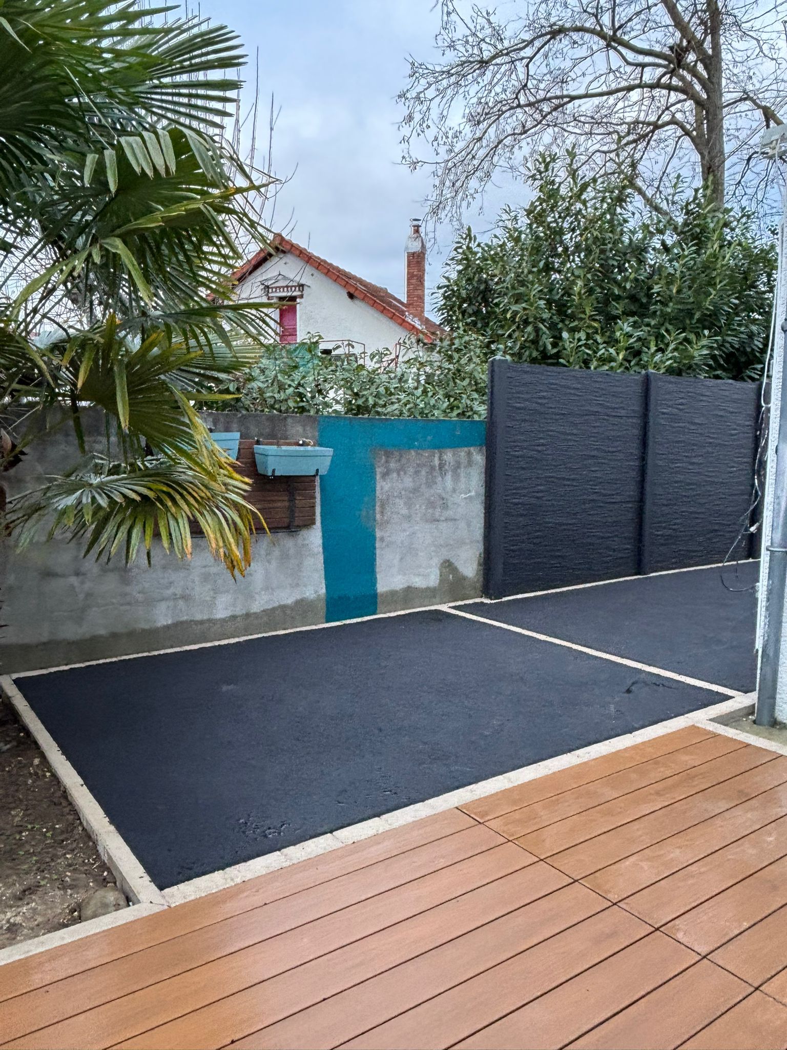 Une allée asphaltée et une terrasse en bois bordent un mur de jardin et une maison.