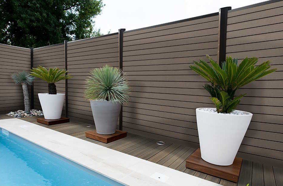 Au bord de la piscine, des plantes en pots blancs sur des plateformes en bois, avec en arrière-plan une clôture à lattes brun foncé.