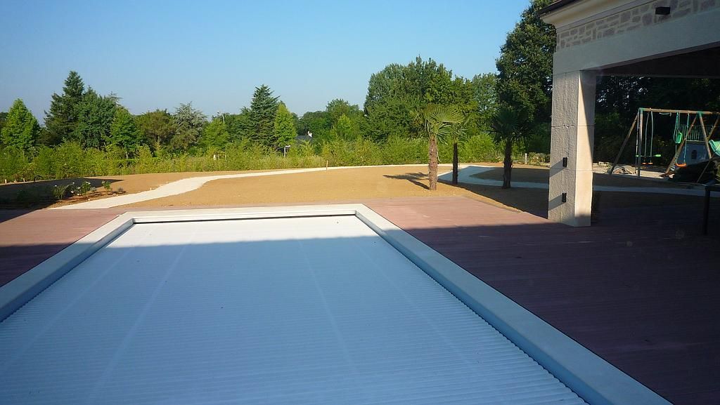 Vue sur une piscine couverte, une terrasse en briques rouges, des arbres et une pelouse. Le ciel est bleu.