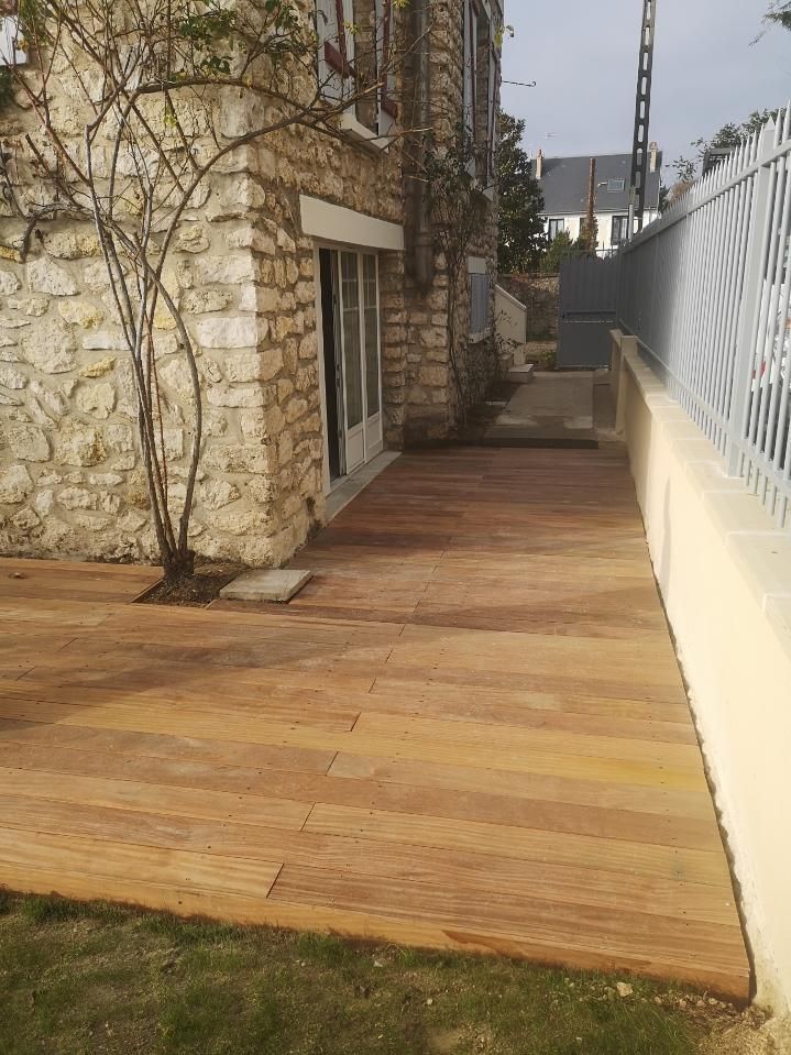 Terrasse en bois jouxtant un bâtiment en pierre et une clôture métallique. Herbe verte au premier plan.