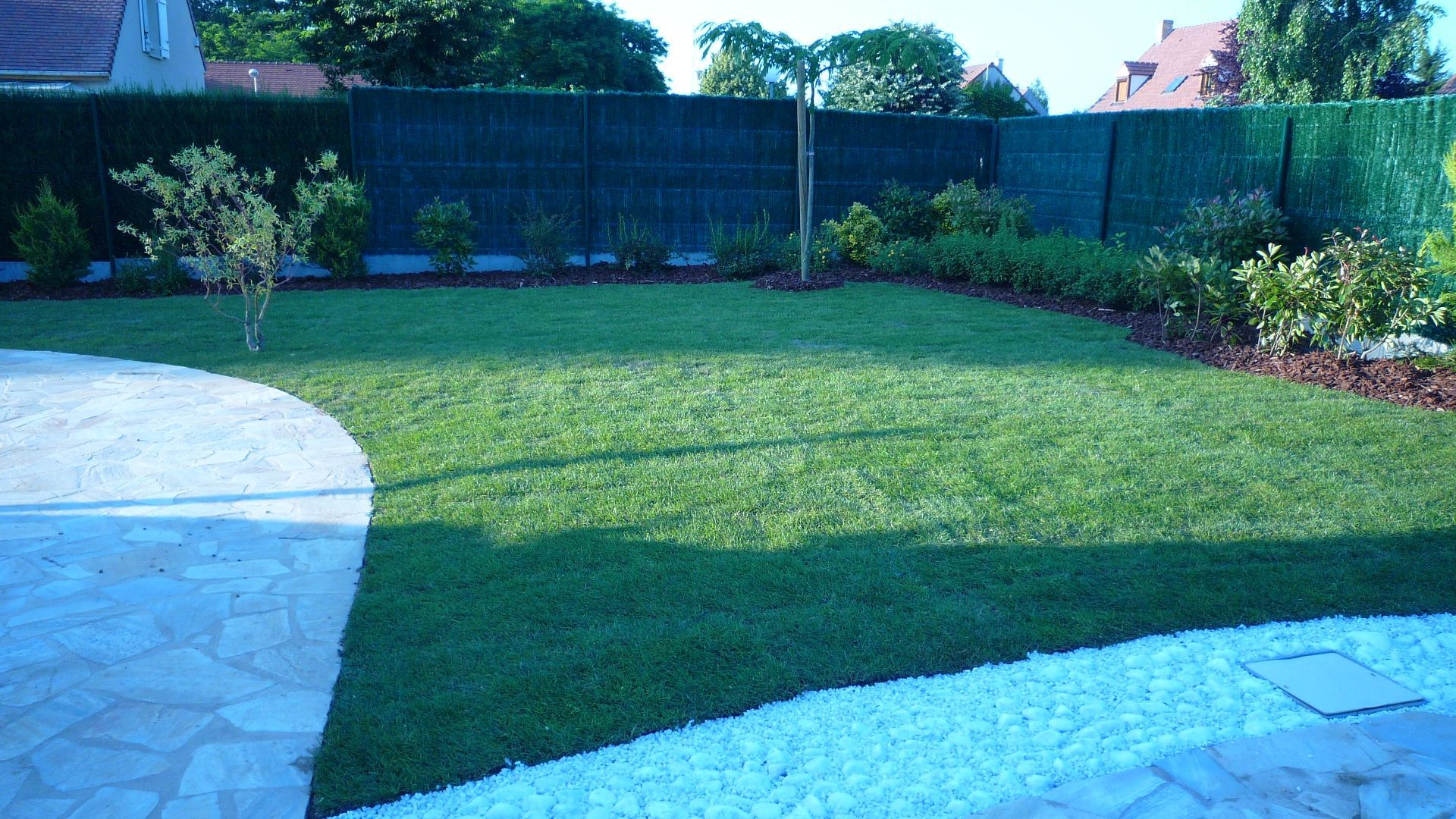Pelouse verte avec un chemin pavé, bordée d'une clôture et d'arbustes.