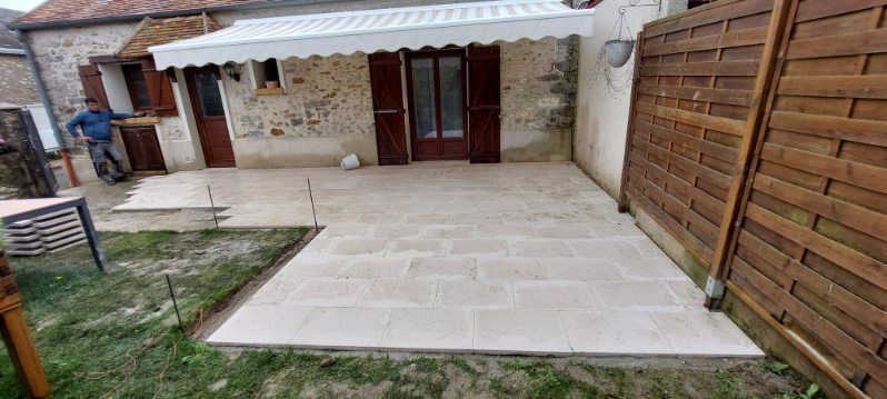 La terrasse est pavée de grands carreaux rectangulaires clairs. Un homme se tient près du bâtiment.