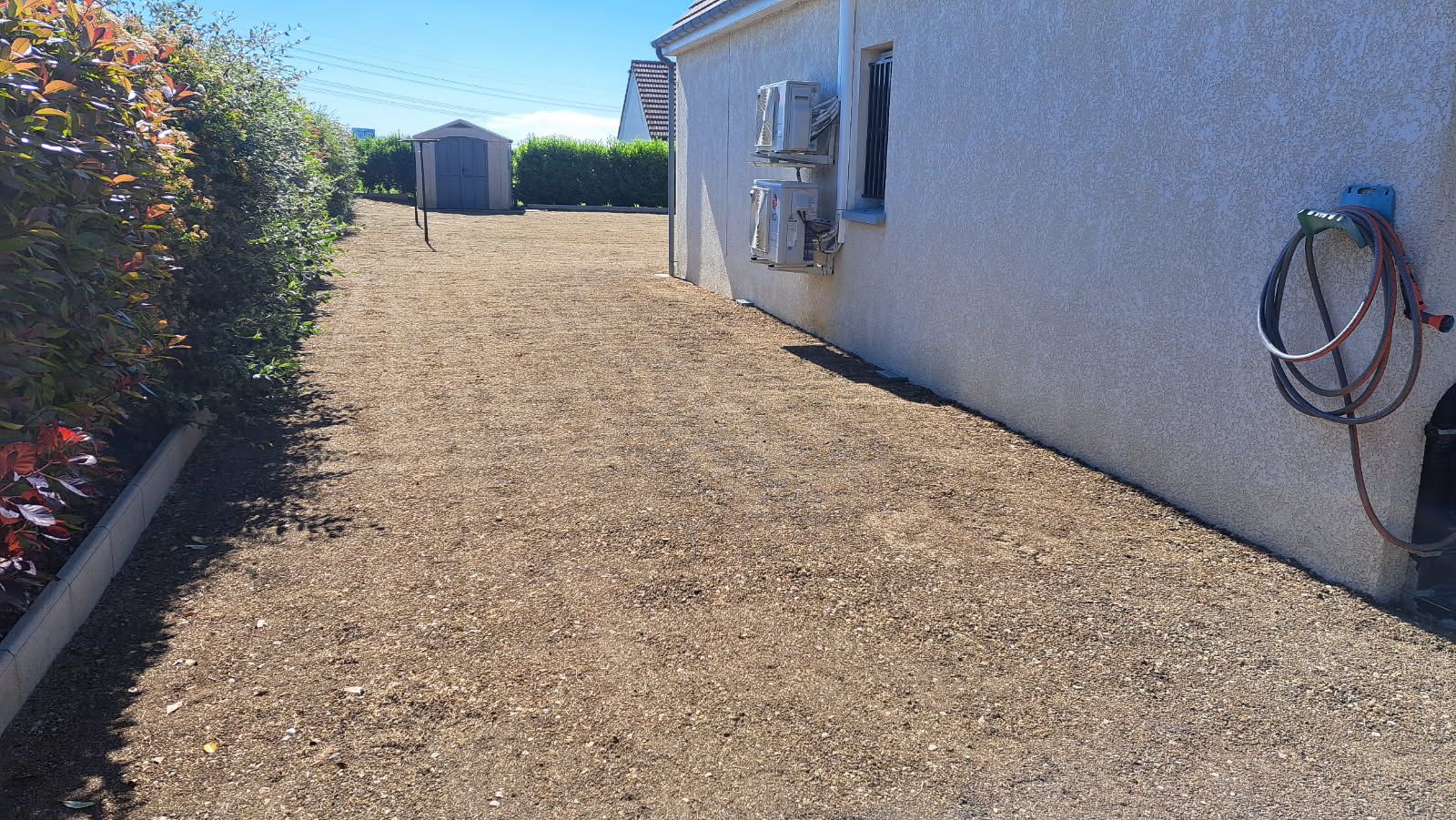 Allée de gravier longeant une maison blanche et des arbustes. Un tuyau d'arrosage est accroché au mur.