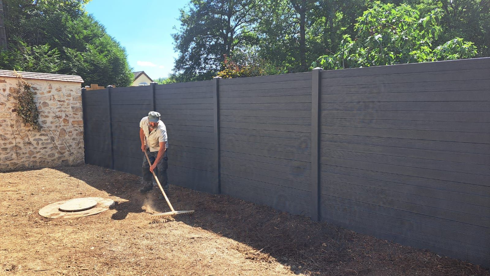 Une personne ratisse du gravier dans un jardin, près d'une clôture gris foncé et d'un mur de pierre, par une journée ensoleillée.
