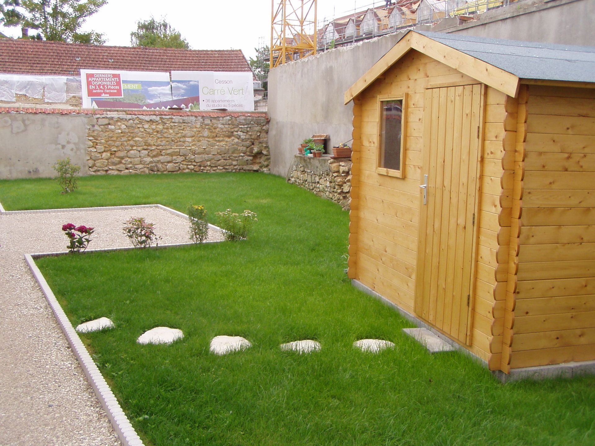 Abri de jardin en bois dans une cour verdoyante avec des pas japonais et des arbustes fleuris ; mur de pierre en arrière-plan.