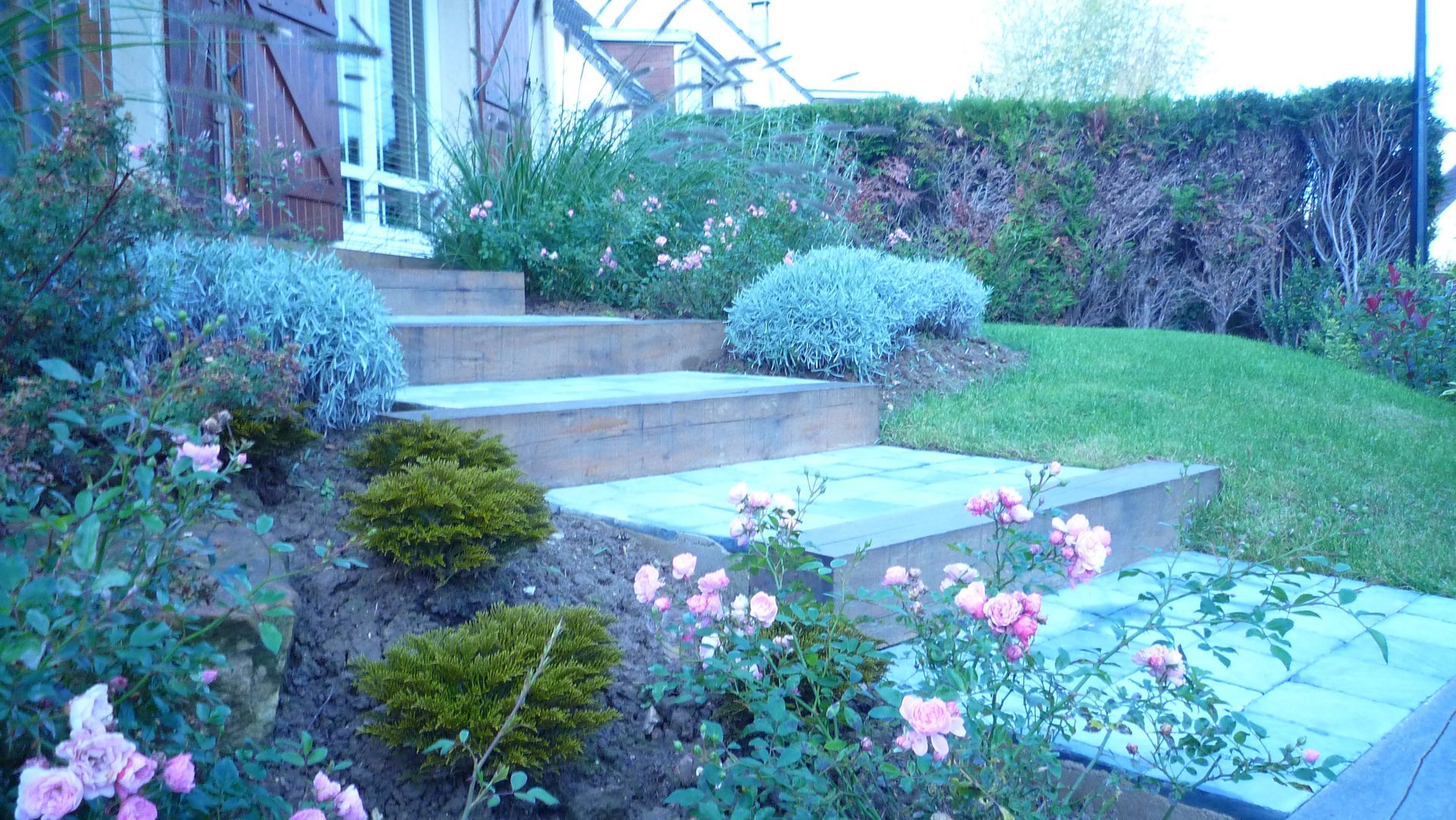 Des marches en pierre mènent à une maison entourée d'un jardin aux buissons verts et aux fleurs roses.
