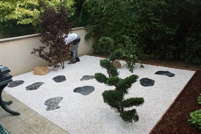 Une personne entretenant un petit jardin zen avec du gravier blanc, des pas japonais et de petits arbres.