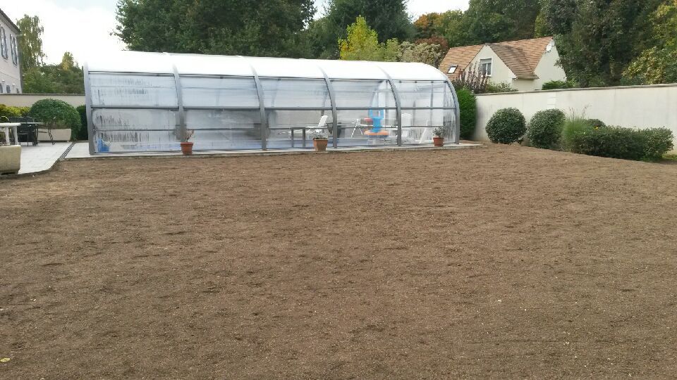 Une serre dans un jardin avec une zone brune nouvellement ensemencée devant.