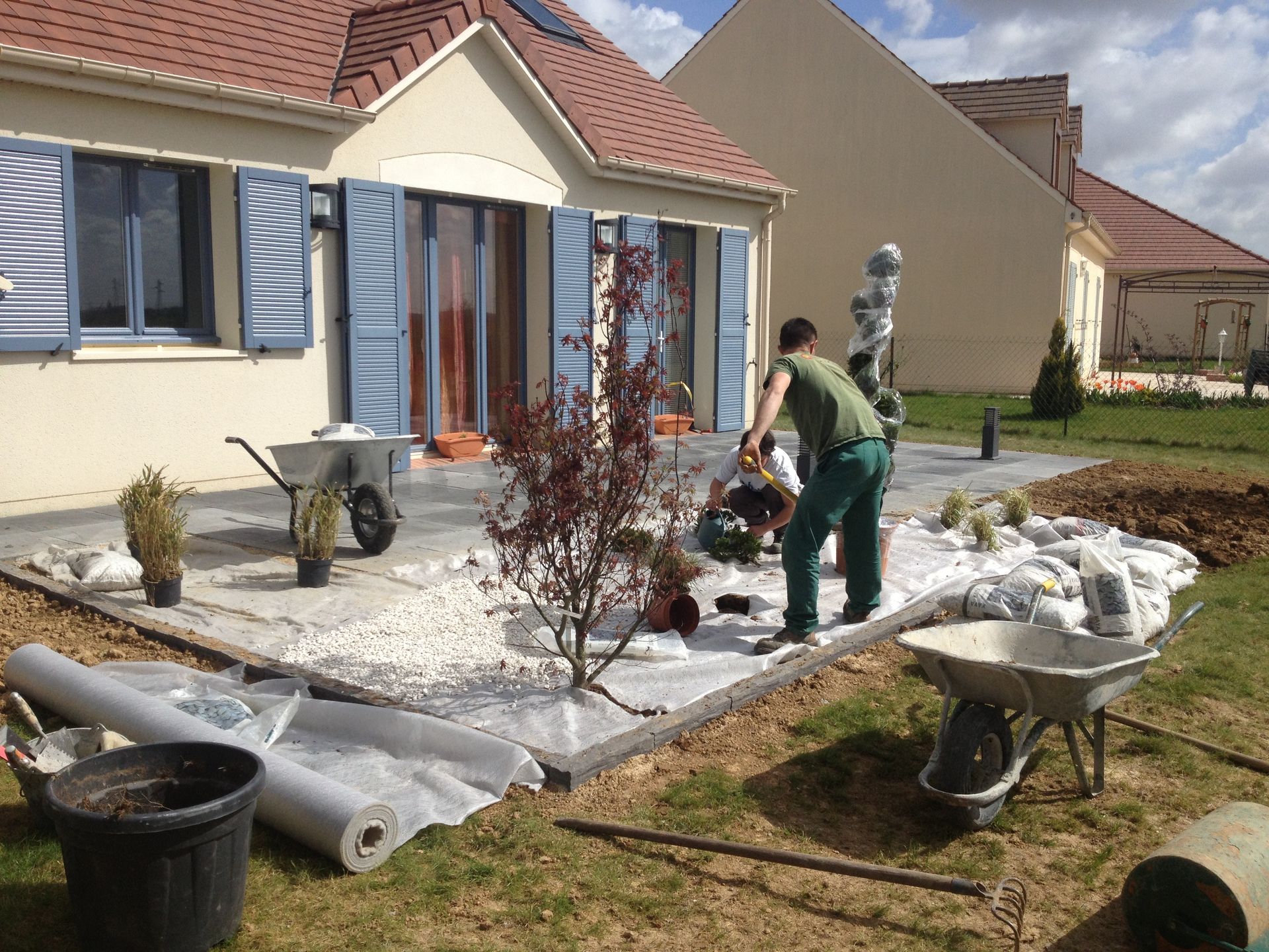 Deux personnes aménagent une terrasse de jardin avec des plantes, des pierres et un petit arbre.