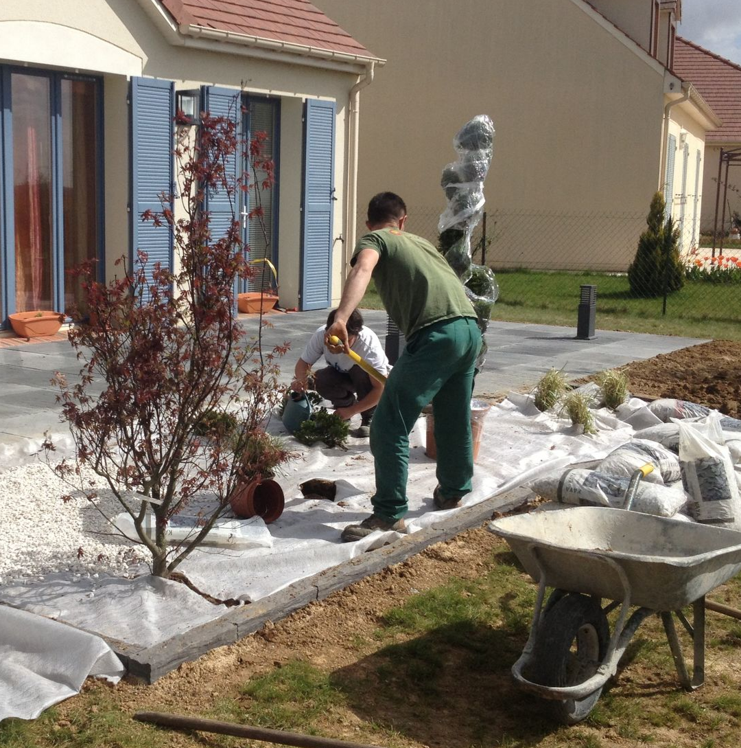 Des ouvriers aménagent un patio avec un petit arbre et des pierres blanches près d'une maison.