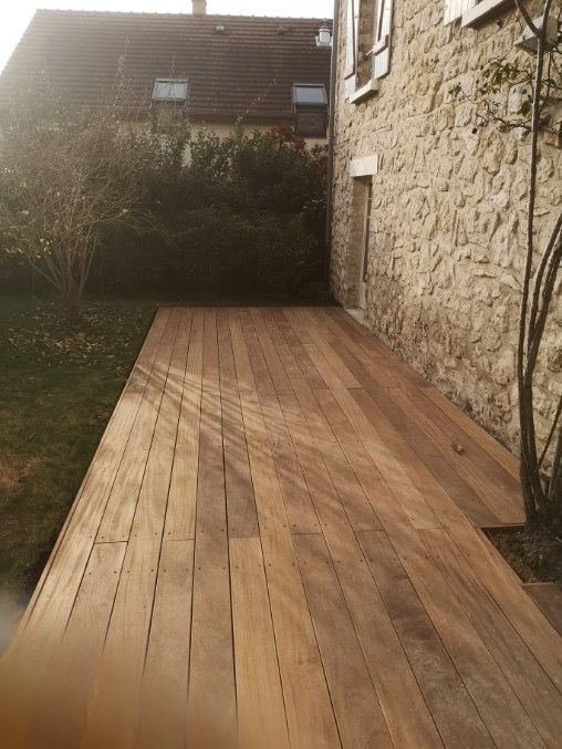 Terrasse en bois jouxtant un bâtiment en pierre ; herbe et buissons en arrière-plan ; journée ensoleillée.