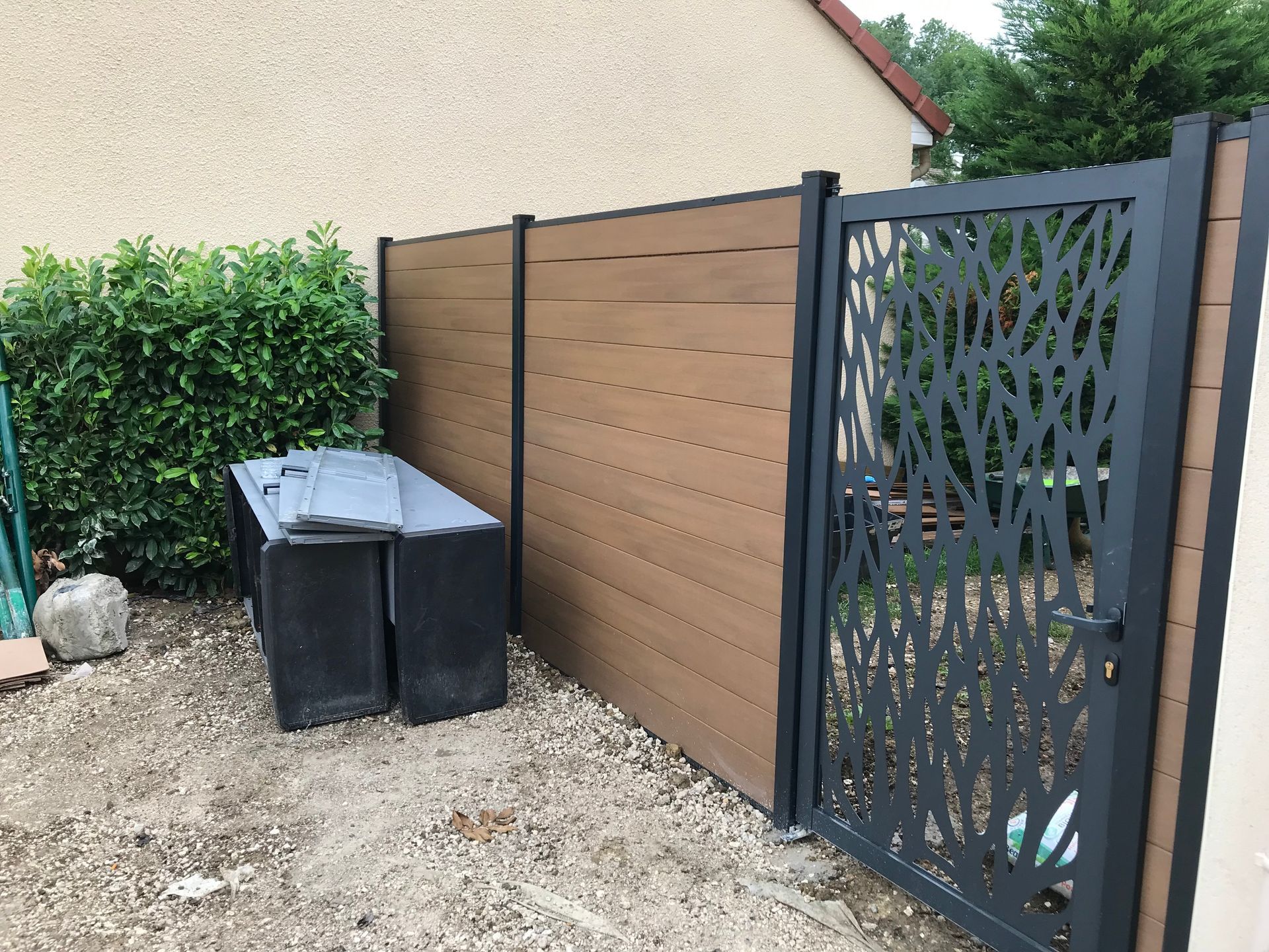 Clôture en bois marron avec un portail décoratif en métal noir, une poubelle noire et un buisson vert.