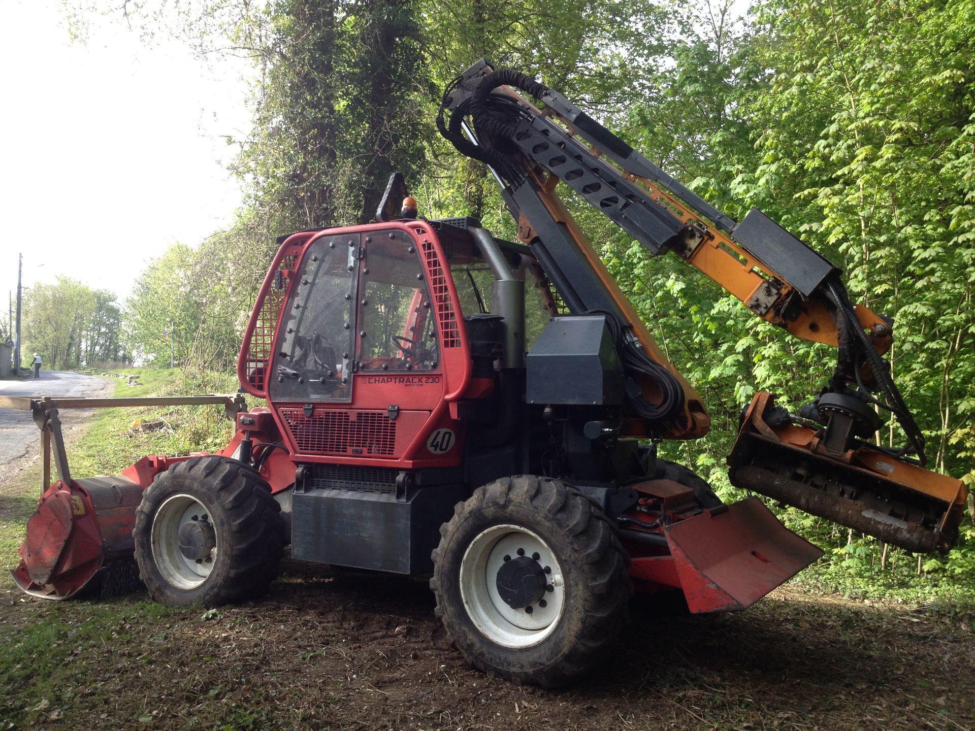 Une machine forestière rouge équipée d'une faucheuse montée sur bras débroussaille une zone boisée.